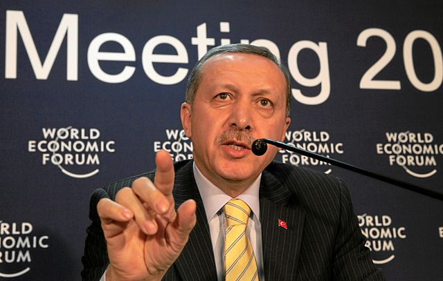 Turkey’s 2009 Prime Minister, Recep Tayyip Erdoğan, raising his finger as he speaks at the microphone at the World Economic Forum in Davos. He is a light brown-skinned man with greying hair and a moustache and wears a grey pinstriped suit, white shirt, yellow tie and red pin. He is in front of a dark blue backdrop with 'Meeting 20...' and 'World Economic Forum' (repeated) in white lettering.