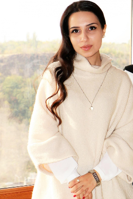 The image shows Ani Kojoyan, a light brown-skinned woman with long wavy brown hair, wearing a white cowl-necked dress and silver chain necklace. She stands in front of a window with a countryside scene faintly visible behind her. Ani's hands are held together in front of her and she is looking straight into the camera with a confident gaze and slight smile.