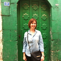 Selin Akyuz, a red-haired woman with a light blue shirt and black shoulder bag, stands in front of a green door and green wall.